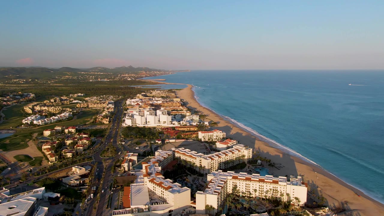The San Jose Del Cabo coastline remains centered from above, with resort roofs, a long sandy beach, calm blue water, distant inland hills, and no visible people.