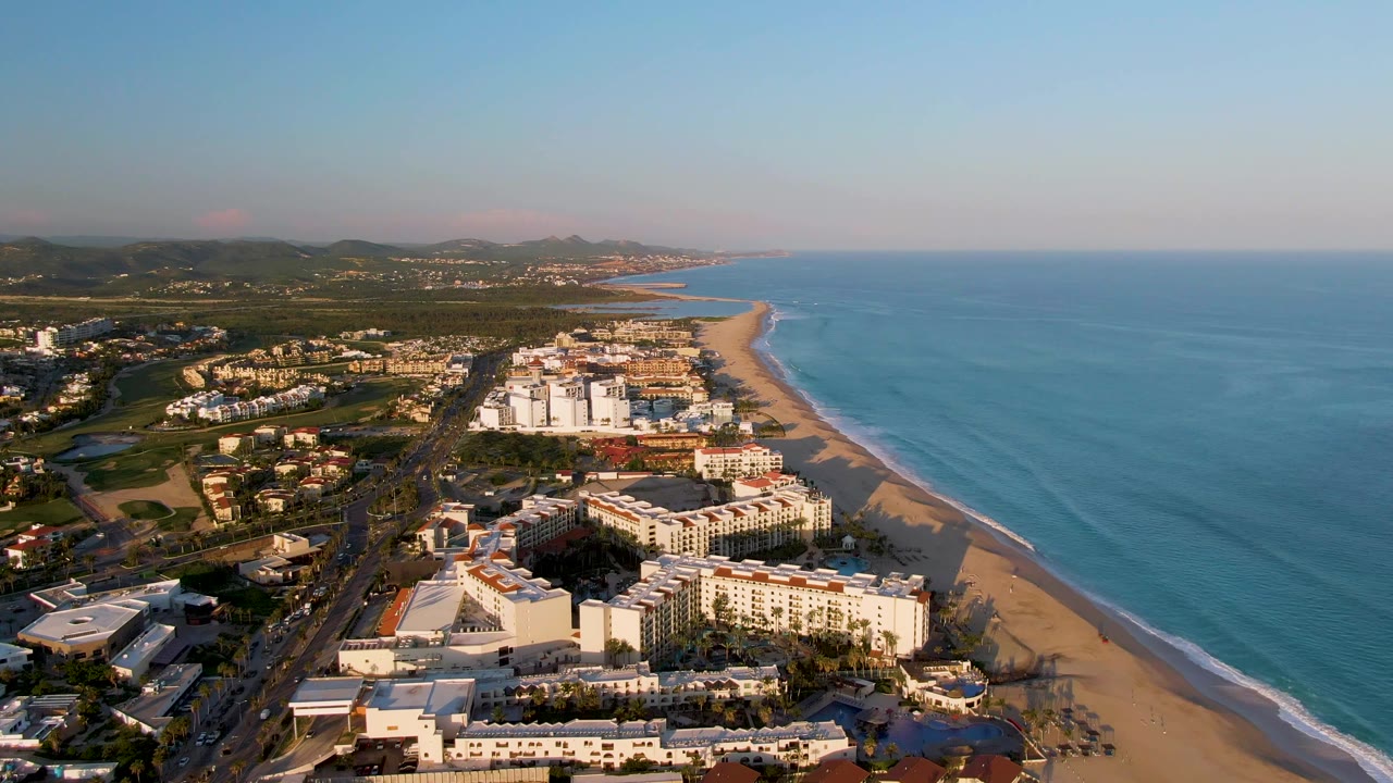 Opening aerial frame over San Jose del Cabo resort buildings, a parallel road, sandy beach, blue ocean, distant hills, and no visible people.