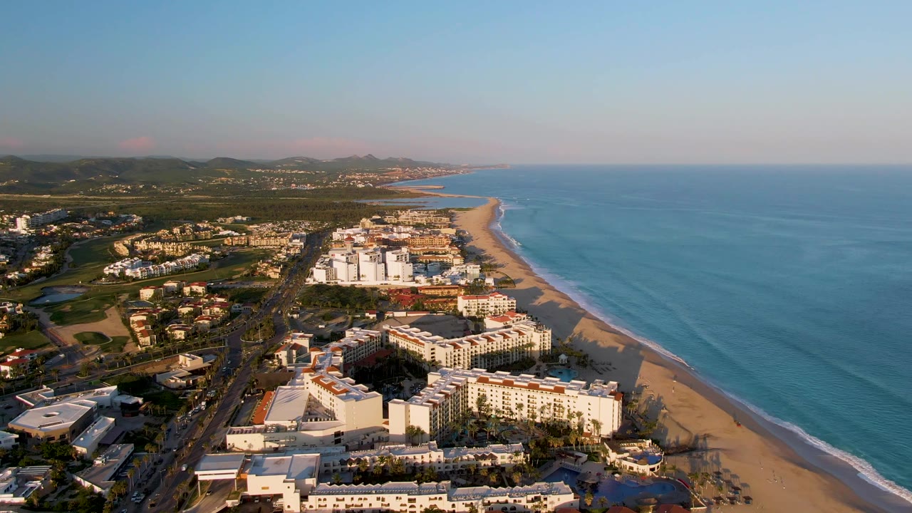 Second aerial frame in San Jose del Cabo showing the resort blocks slightly farther along the shore, with surf lines on the beach, road traffic at left, and no visible people.
