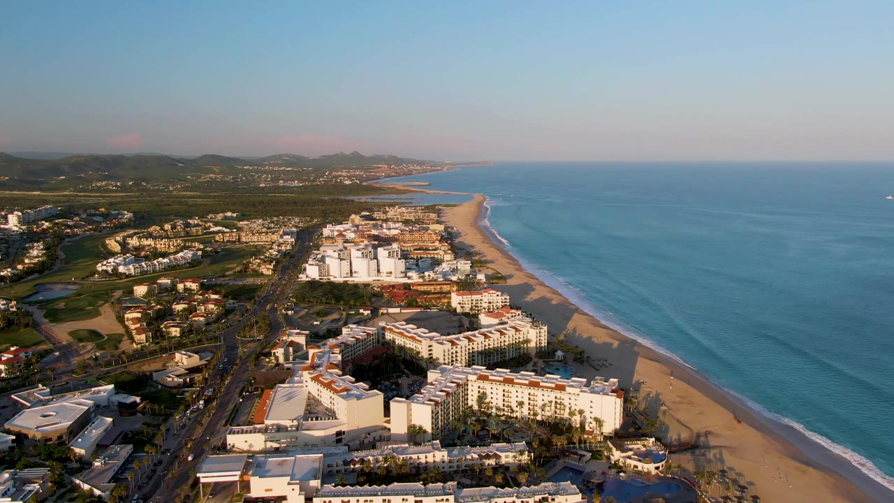 Third aerial frame over San Jose del Cabo with white beachfront buildings centered, the ocean widening to the right, distant coastline ahead, and no visible people.