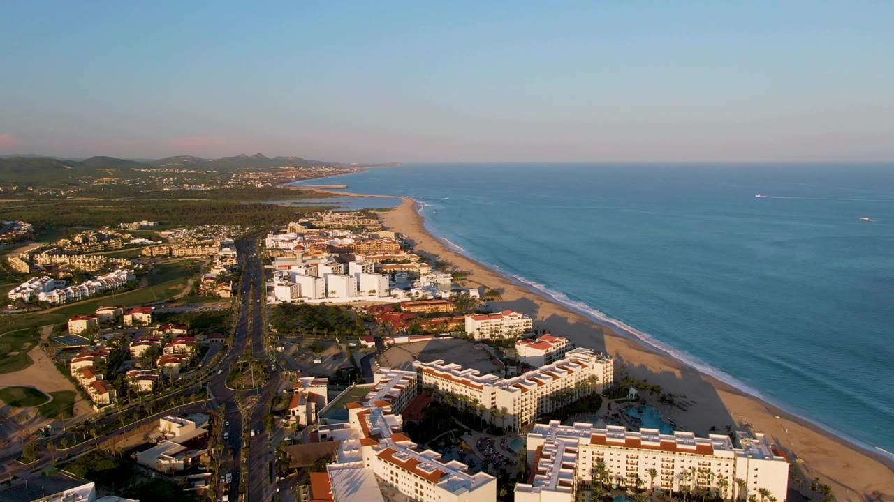 Opening aerial frame over San Jose Del Cabo shows beachfront buildings, a straight coastal road, sandy shore, blue ocean, distant hills, clear sky, and no visible people.
