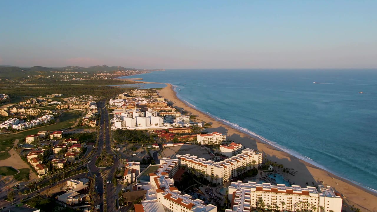 One second into the San Jose Del Cabo aerial, the coastal road sits near center with white beachfront buildings below, ocean to the right, hills beyond, and no visible people.