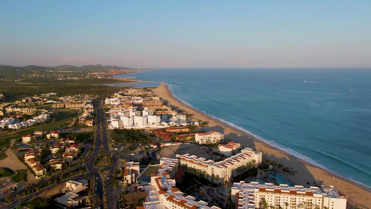 Mid-clip aerial frame in San Jose Del Cabo shows the beach running diagonally beside blue water, with resort-like buildings and a road in warm light and no visible people.