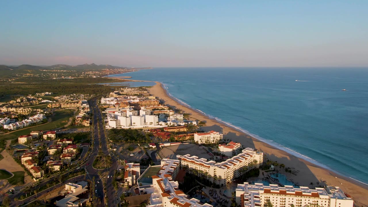 Aerial view looking along the beach with buildings, road, ocean, and hills.