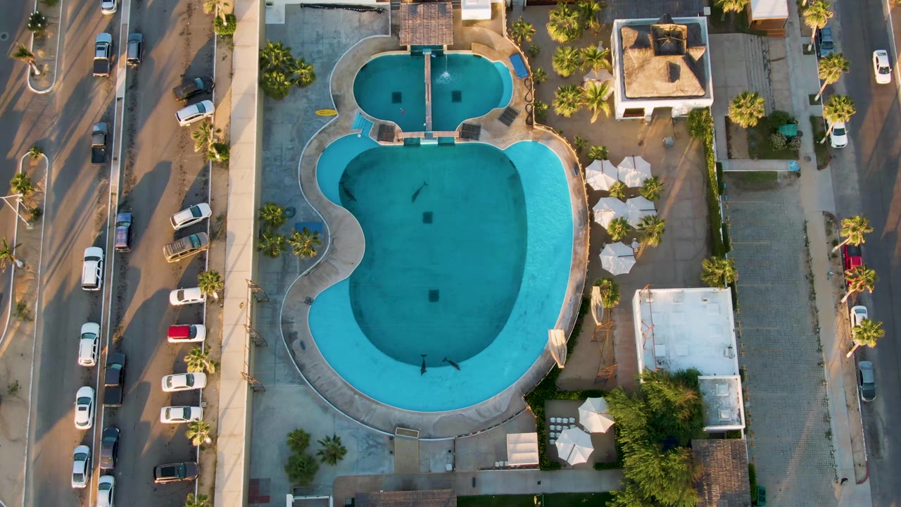 Third San Jose del Cabo frame from above, with the pool centered, patio umbrellas and small roofs to the right, parked vehicles on the street, and no visible people.