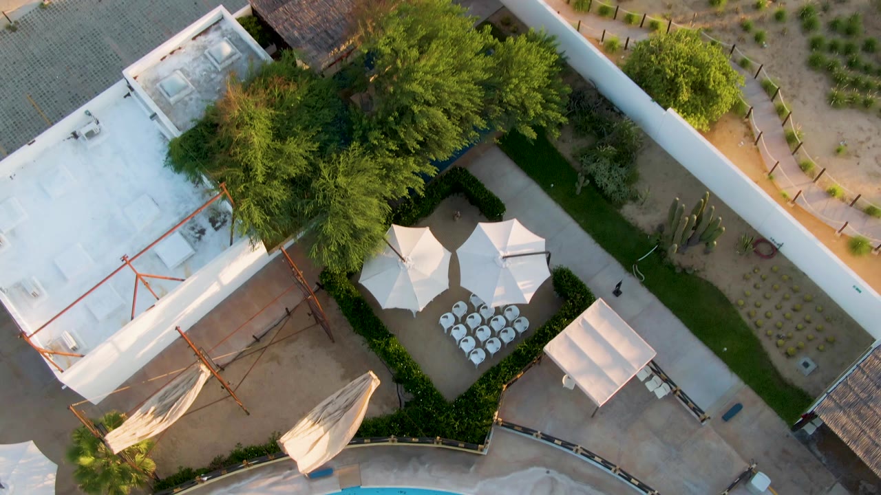 Top-down aerial frame in San Jose del Cabo showing a white building roof, courtyard umbrellas, green trees, desert landscaping, and surrounding walls, with no visible people.