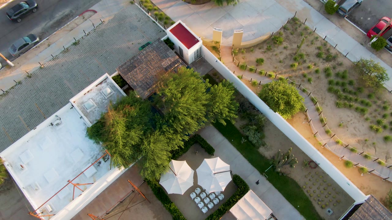 Overhead San Jose del Cabo view with the courtyard shifted lower in frame, revealing more street pavement, parked cars, sidewalks, and planted landscaping, with no visible people.