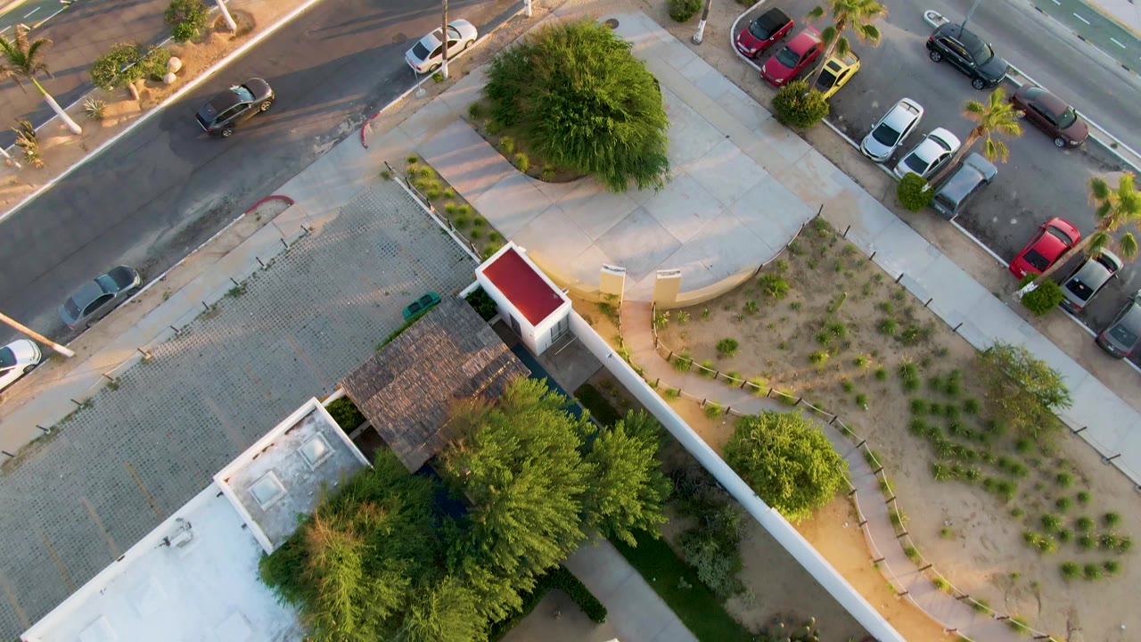 Aerial San Jose del Cabo frame showing a street corner, parked cars, palm trees, a curved sidewalk, sparse desert planting, and the courtyard wall at the lower edge, with no visible people.