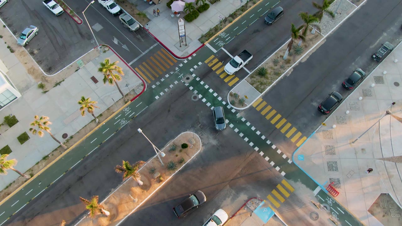 Overhead aerial frame in San Jose Del Cabo showing a diagonal street intersection, green bike lanes, yellow crosswalks, parked cars, palm trees, and no visible people.