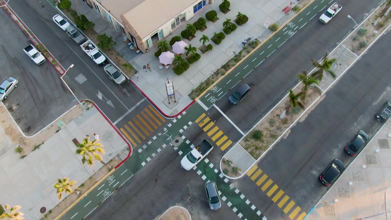 Aerial frame in San Jose Del Cabo with the intersection centered, low-rise buildings at the top edge, parked cars along the curb, palm trees, and no visible people.