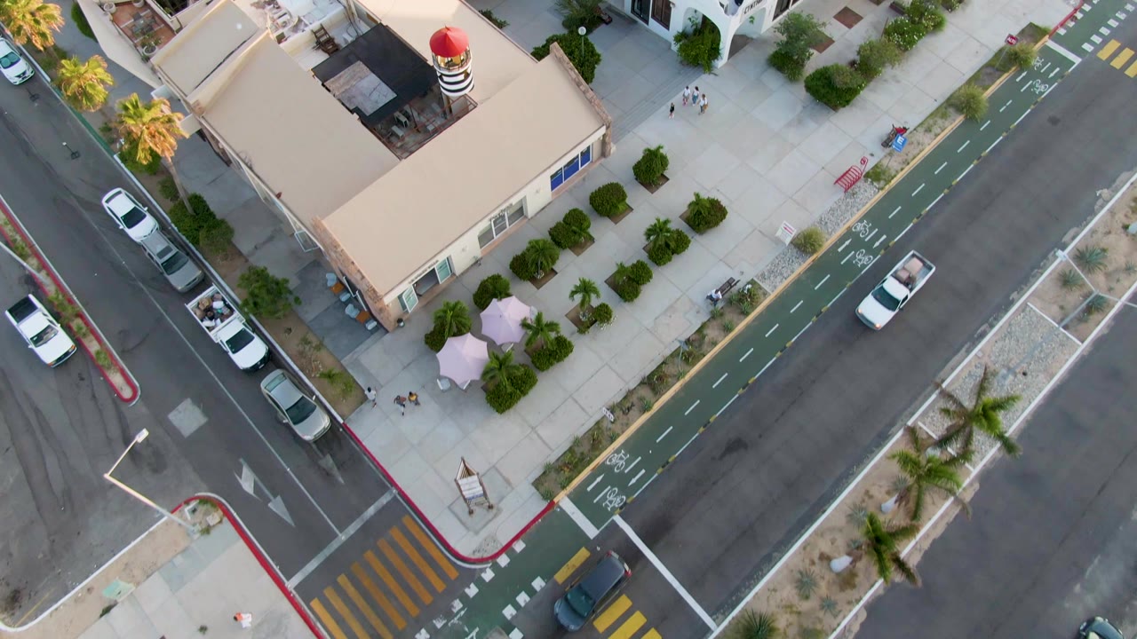 Top-down San Jose Del Cabo frame showing tan and white buildings around a marked intersection, with cars on the road, palm trees along sidewalks, and no visible people.
