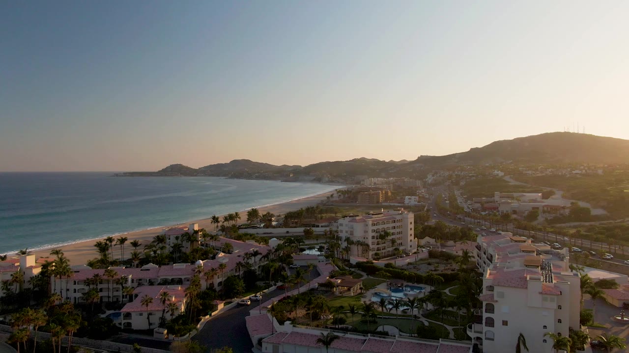 A high aerial view over San Jose del Cabo shows the ocean and sandy beach on the left, resort buildings and palms below, hills in the distance, and no visible people.