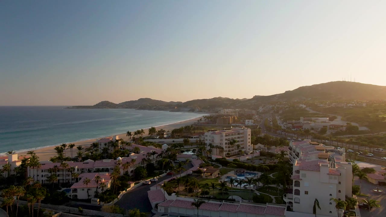 The San Jose del Cabo coastline remains in a wide aerial view, with blue water and beach at left, palm-lined resort grounds below, hazy hills beyond, and no visible people.