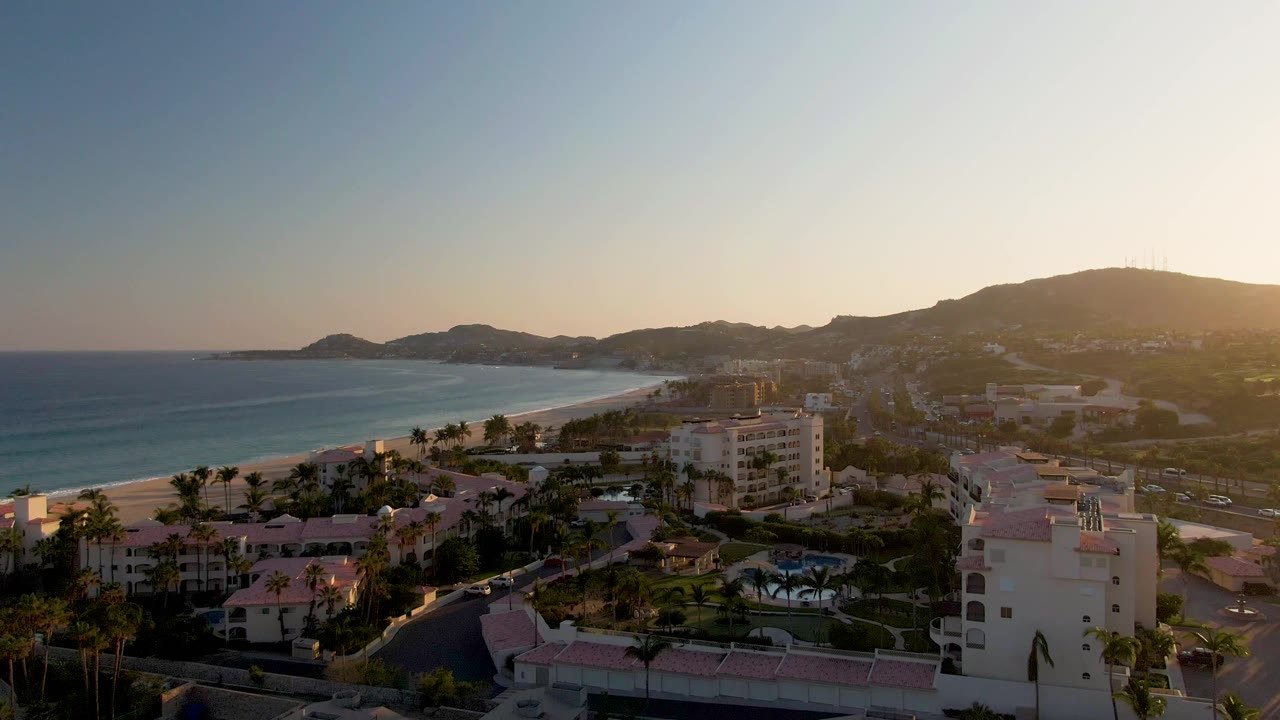 A slightly closer aerial angle over San Jose del Cabo shows the curving beach, ocean, resort blocks, palms, hill silhouettes, golden light from the right, and no visible people.