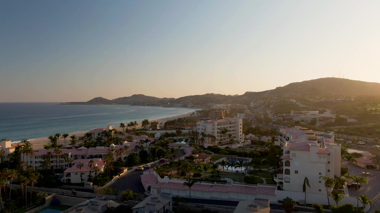 Lower aerial view of resort buildings, palms, beach, ocean, and distant hills.
