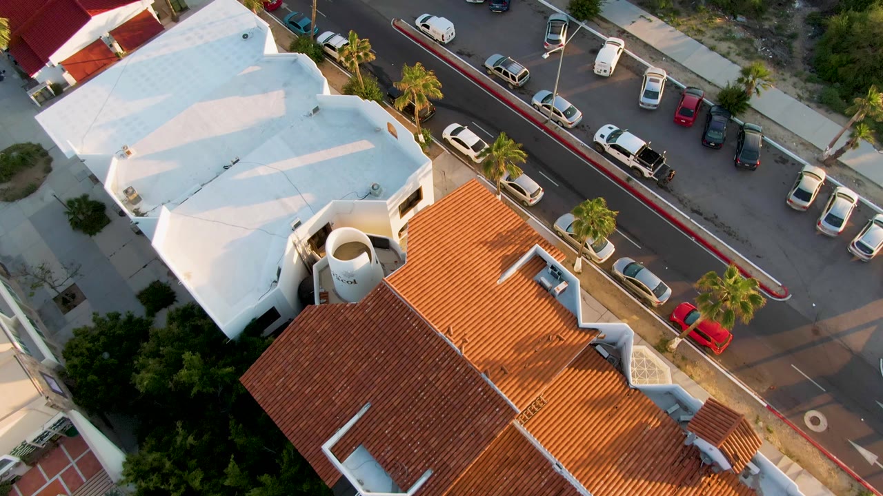 Overhead frame in San Jose del Cabo showing a white-roof building, a red tile roof, curbside parked cars, palm trees, and no visible people.