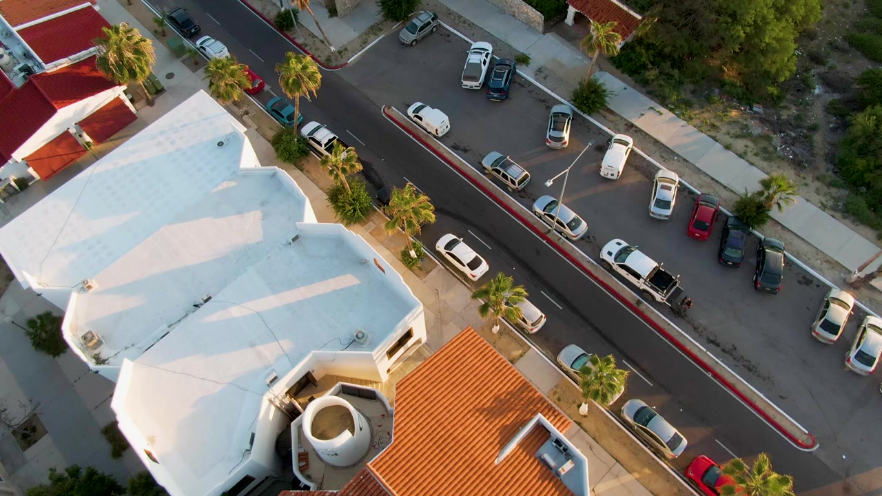 Aerial view over a white-roof building beside a street with parked cars and palms.