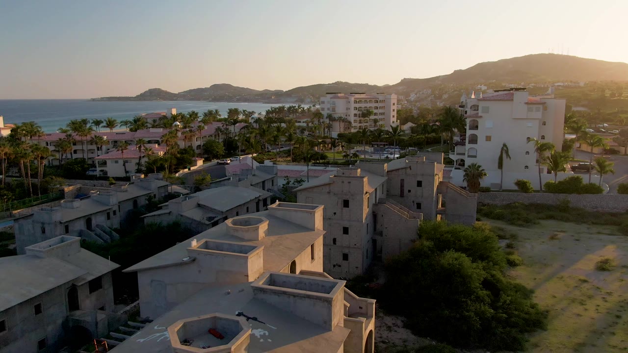 Aerial frame over residential buildings in San Jose Del Cabo, with palm trees, the ocean, distant hills, warm sunset light, and no visible people.