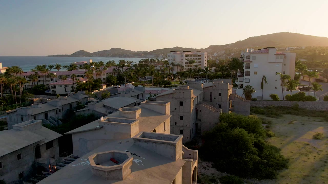 Drone view of coastal homes and palms with ocean and hills in warm light.