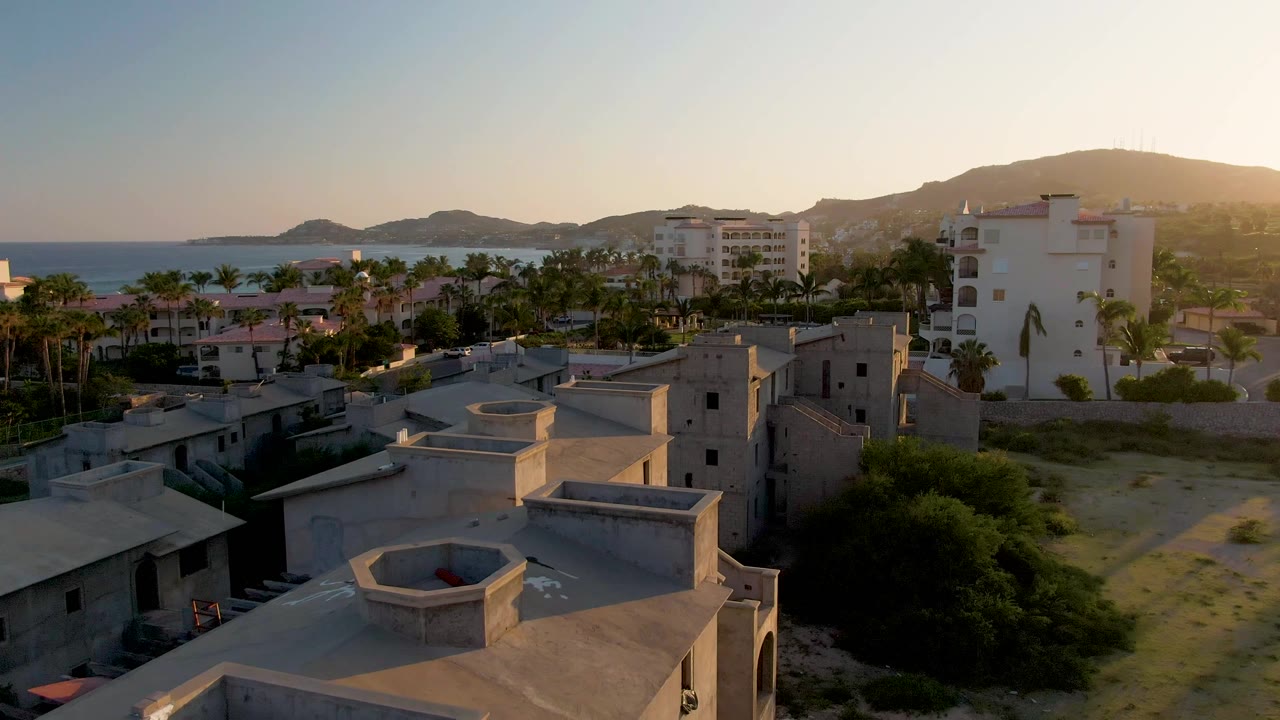 Mid-clip aerial view in San Jose Del Cabo showing closer flat rooftops, palm-lined buildings, the ocean to the left, hills in the distance, and no visible people.