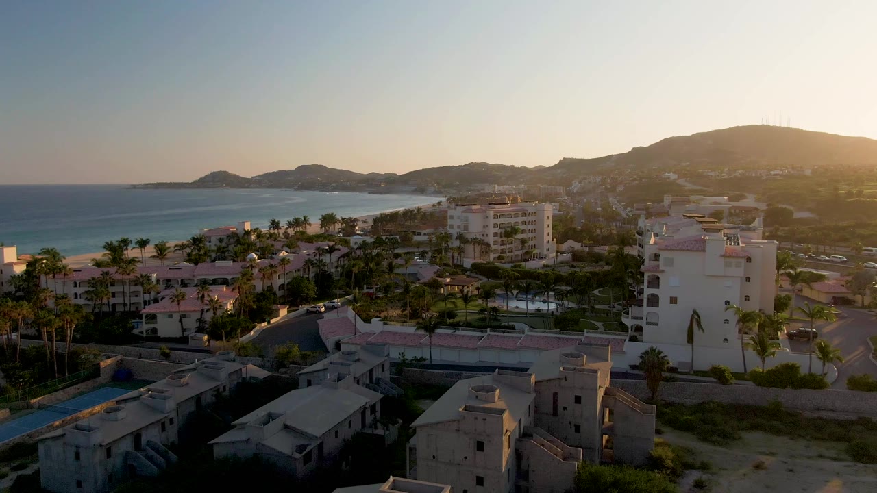 Aerial view over San Jose Del Cabo residential buildings, palm trees, ocean, and distant hills in warm sunset light, with no visible people.