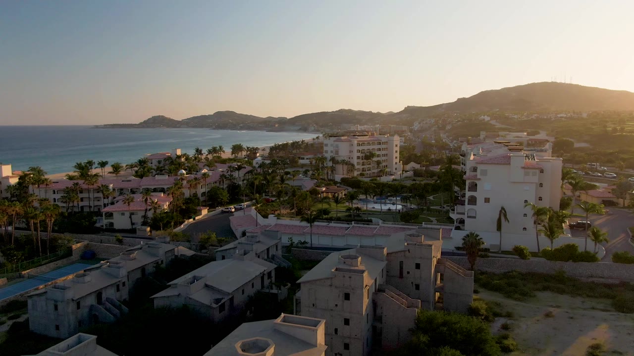 San Jose Del Cabo rooftops and palm trees fill the foreground, with blue ocean to the left and hazy hills behind the buildings, with no visible people.