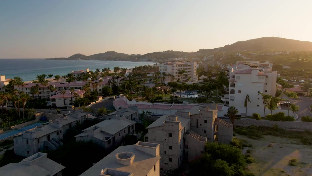 Aerial composition in San Jose Del Cabo showing a prominent white building, clustered palms, nearby homes, the ocean, and hills under golden light, with no visible people.