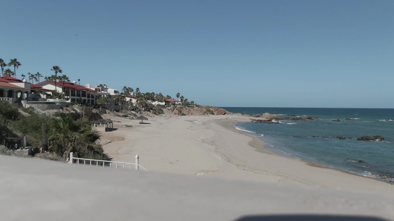 Opening view of Punta Pescadero beach with a pale foreground edge, red-roofed buildings at left, gentle surf at right, rocky shoreline ahead, and no visible people.
