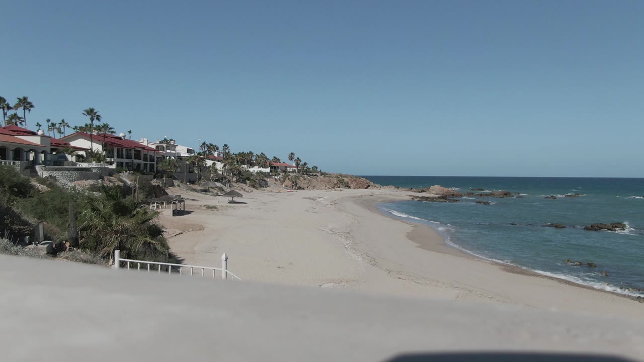 Wide Punta Pescadero beach view at the start of the clip, with pale sand, blue water, red-roof buildings on the left bluff, palm trees, and no visible people.