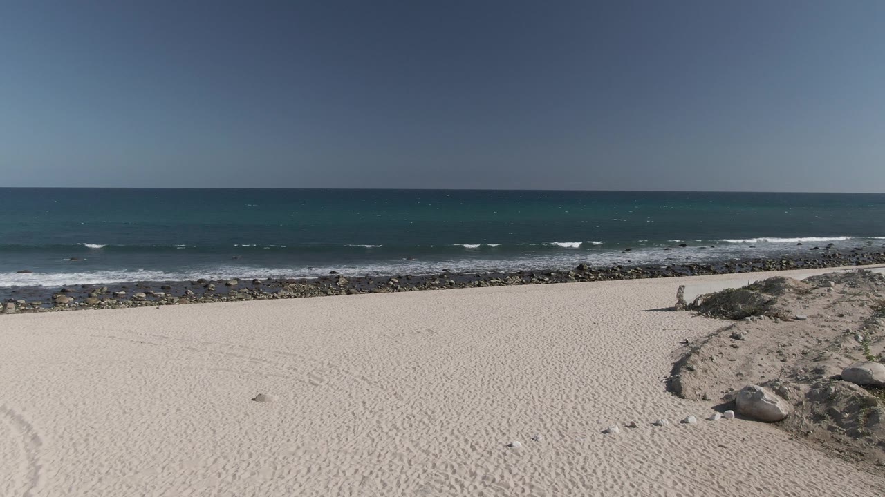 Costa Azul Los Cabos shoreline frame with broad pale sand, scattered footprints, a dark rock strip, small white wave crests, and no visible people.