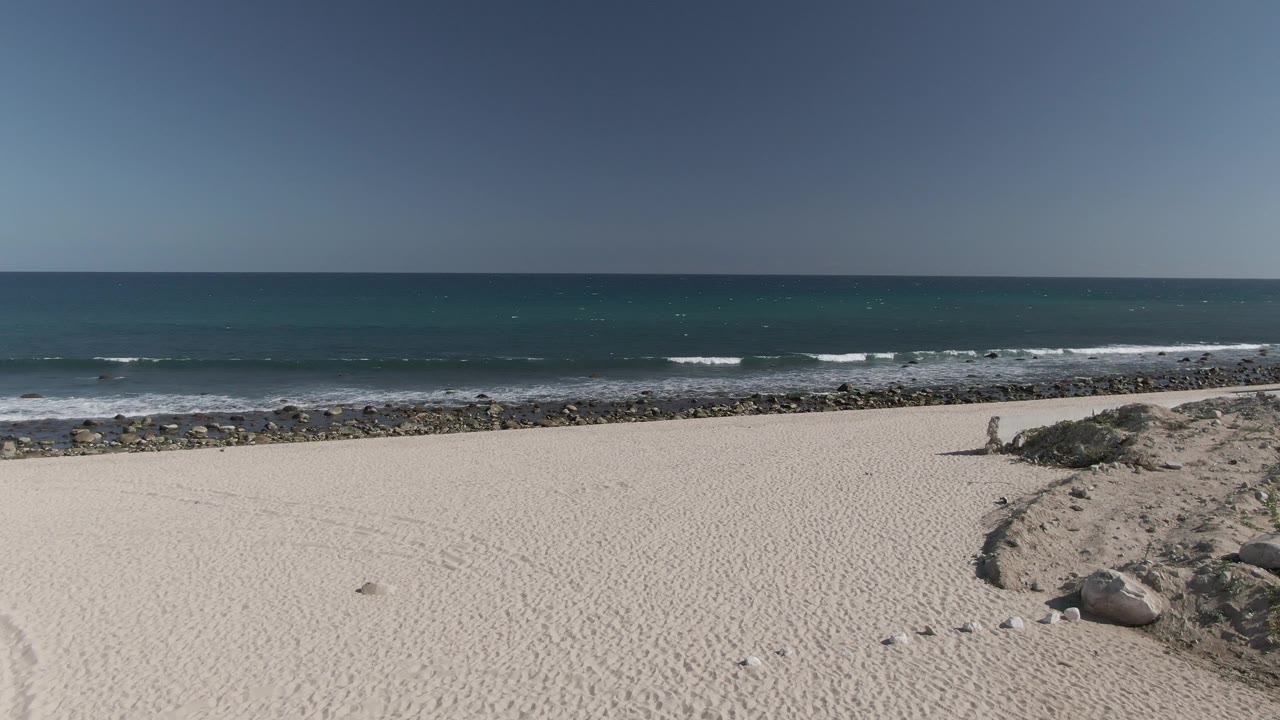 Elevated Costa Azul Los Cabos beach view where white foam advances near the rocky shoreline, with open sand and no visible people.