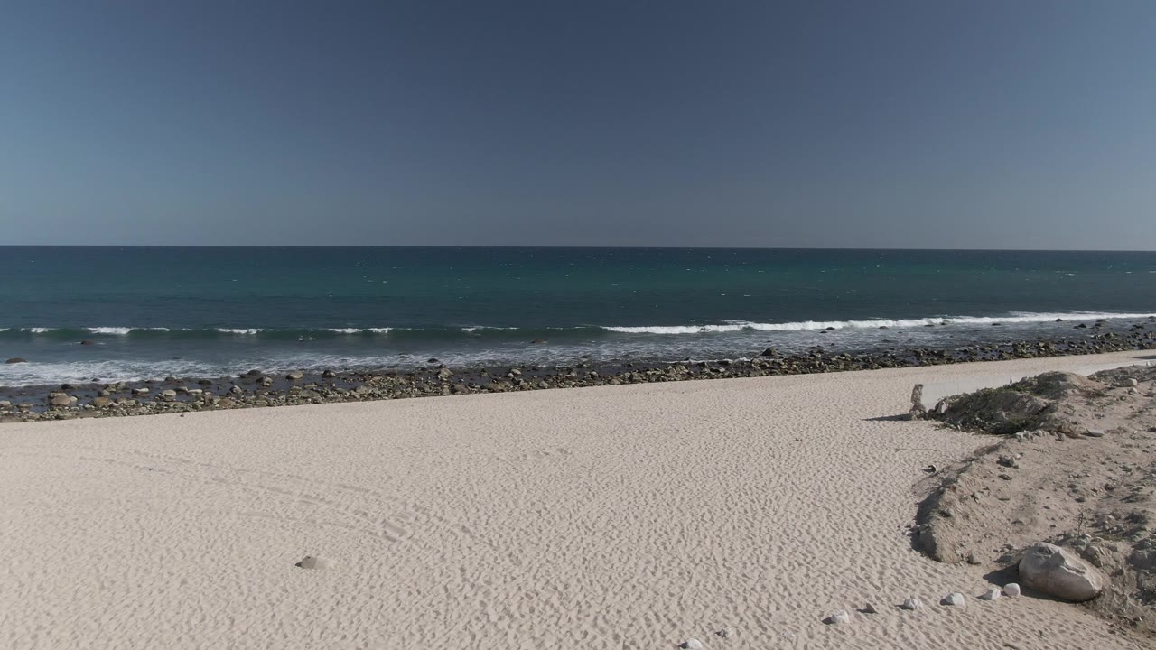 Costa Azul Los Cabos beach frame with a low sandy ridge at right, pale open sand, rocks along the shore, rolling ocean waves, and no visible people.