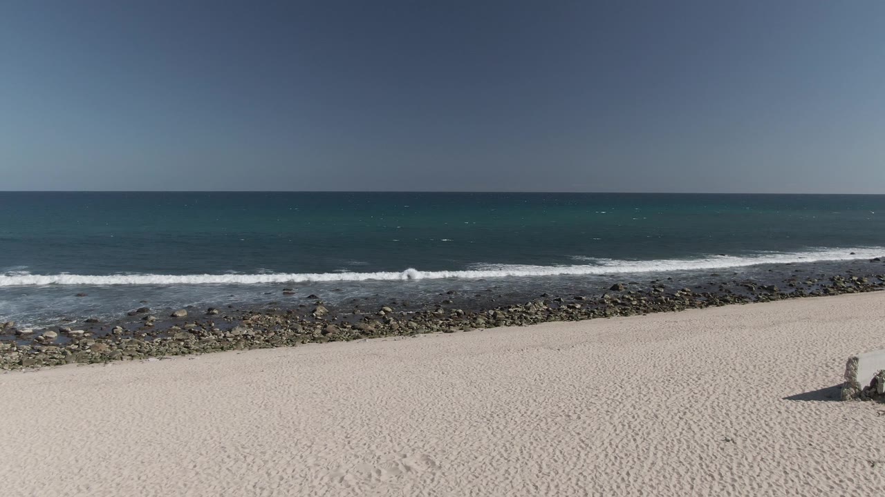 Aerial frame of Costa Azul Los Cabos showing pale sand in the foreground, a rocky shoreline, white surf, blue ocean, hazy sky, and no visible people.