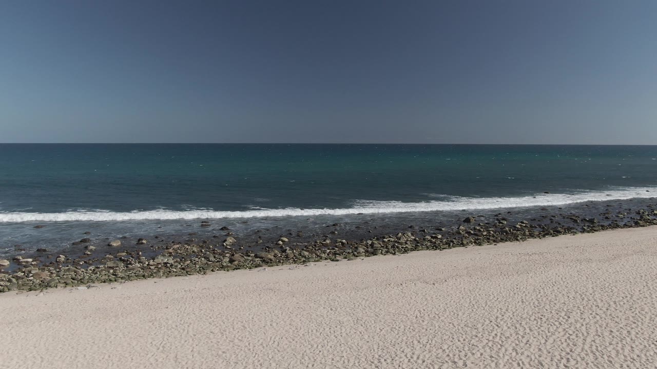 Aerial view at Costa Azul Los Cabos with the white beach lower in frame, a dark rock band at the shore, thin white surf, open ocean, and no visible people.