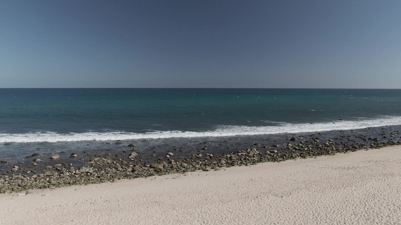 Costa Azul Los Cabos shoreline frame with more rocky beach visible at the waterline, white waves pushing toward pale sand, blue-green ocean, and no visible people.