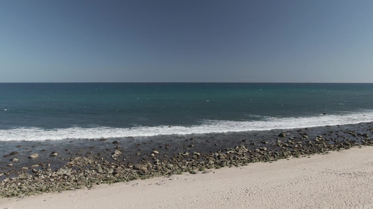 Aerial coastal view with rocks across the foreground and waves offshore.