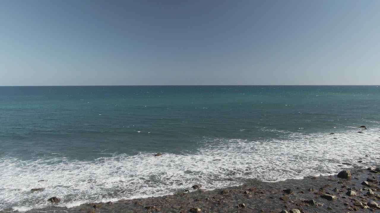 At Costa Azul Los Cabos, a wider band of white foam covers the rocky shoreline while darker blue water sits beyond the turquoise shallows, with no visible people.
