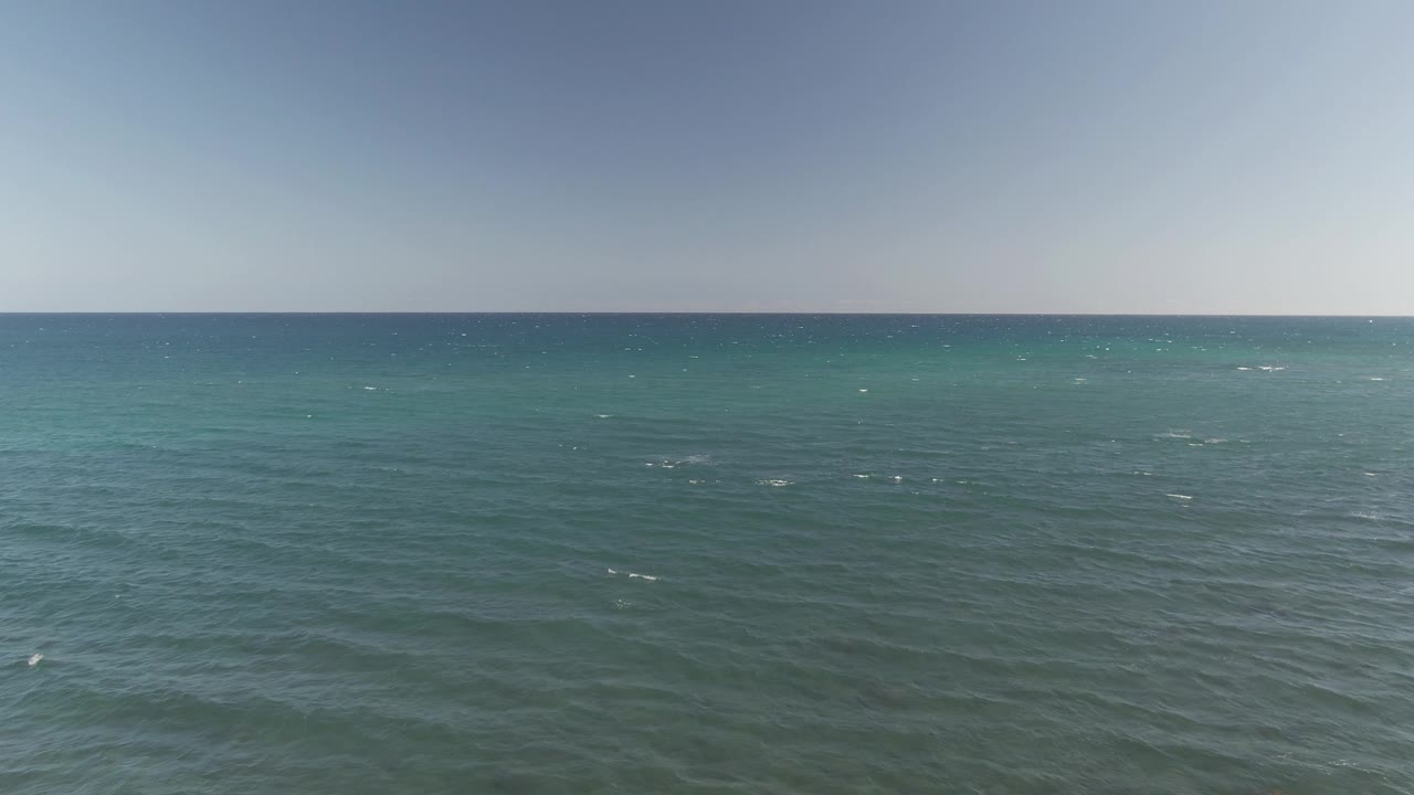 The Costa Azul Los Cabos ocean appears slightly darker in the foreground, with low wave ridges, a turquoise strip near the horizon, clear sky, and no visible people.
