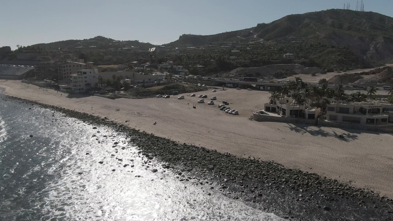 Aerial frame from Costa Azul Los Cabos showing bright ocean glare at left, a dark rock strip along the sand, beachfront buildings, parked cars, hillside terrain, and no visible people.