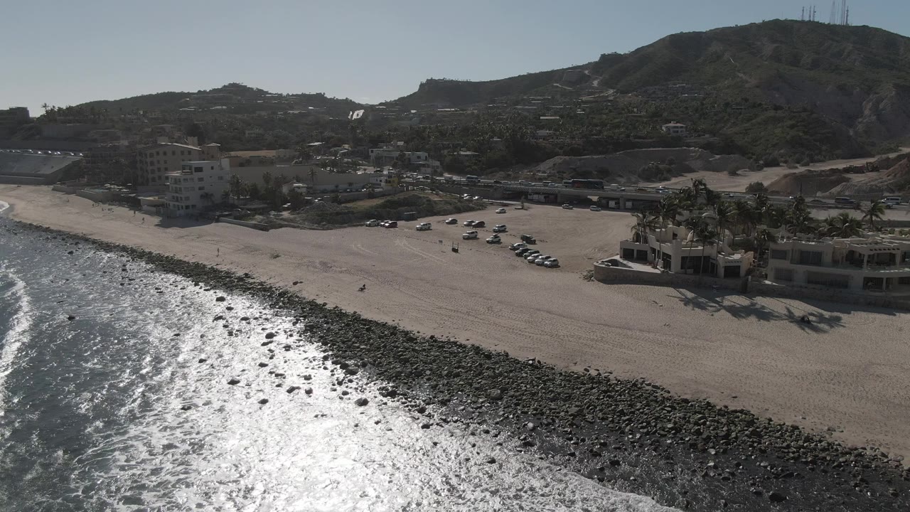 Wide aerial beach view with reflective water, rocks, sand, cars, buildings, and hills.
