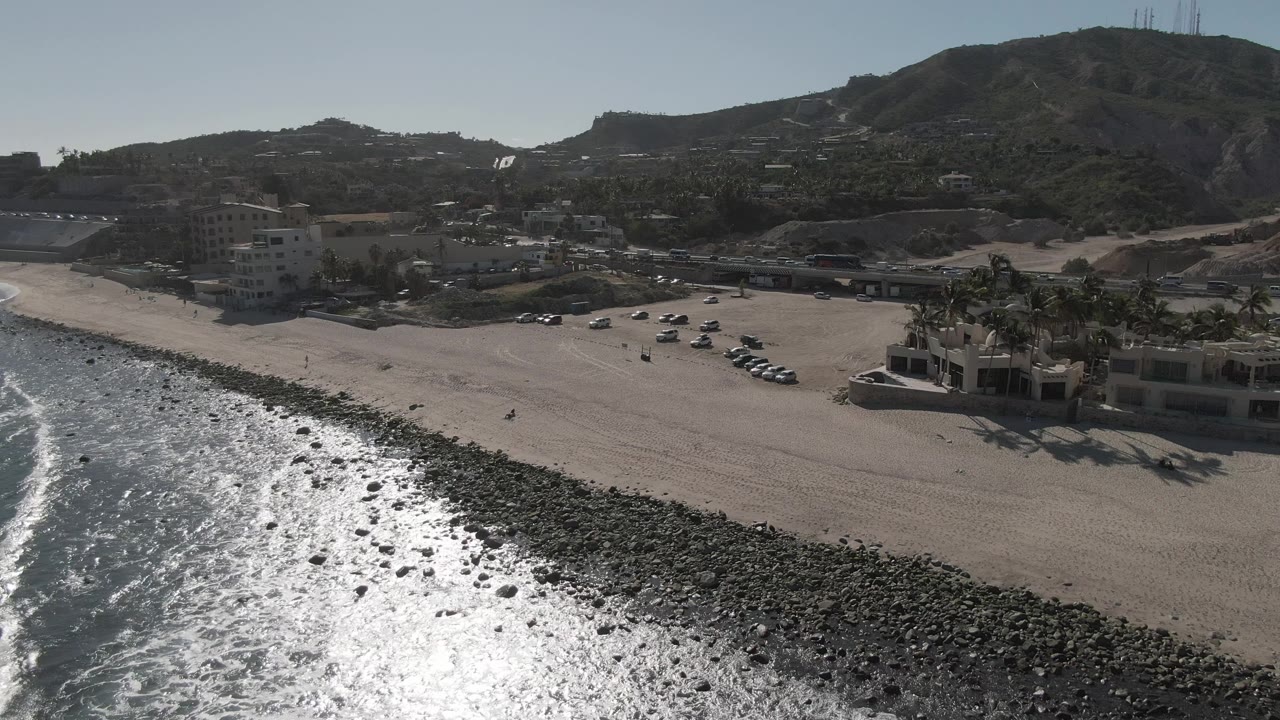 Aerial view over Costa Azul Los Cabos with white wave foam beside the rocky shore, a broad sandy beach, cars lined near the beach access area, buildings, palms, and no visible people.