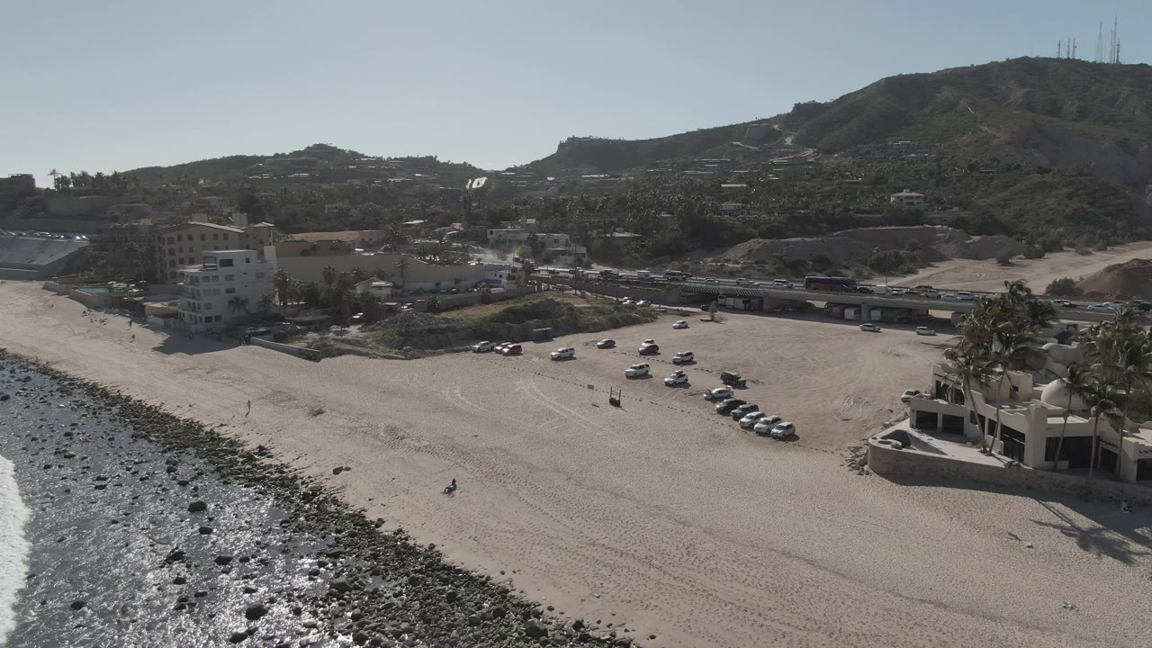 Aerial view over Costa Azul Los Cabos showing the rocky waterline at left, open sand, parked cars, nearby buildings, a coastal road, hillside terrain, and no visible people.