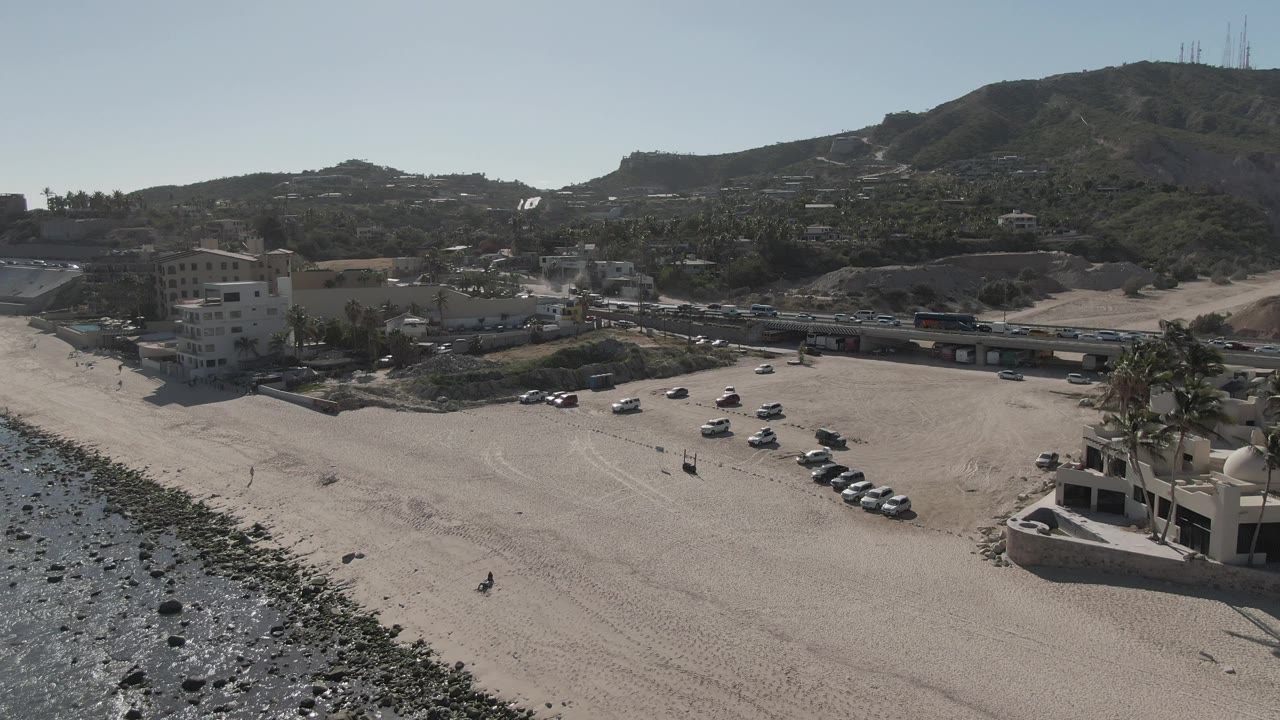 Drone view of beach parking, coastal road, buildings, rocky shore, and hills.