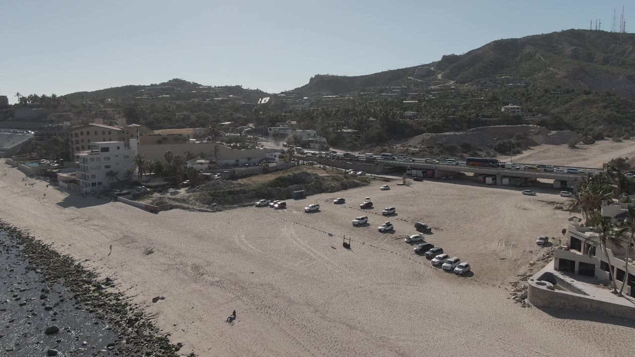 Aerial frame over Costa Azul Los Cabos showing parked vehicles grouped on the sand, the rocky shore along the lower left, road traffic, hillside buildings, and no visible people.