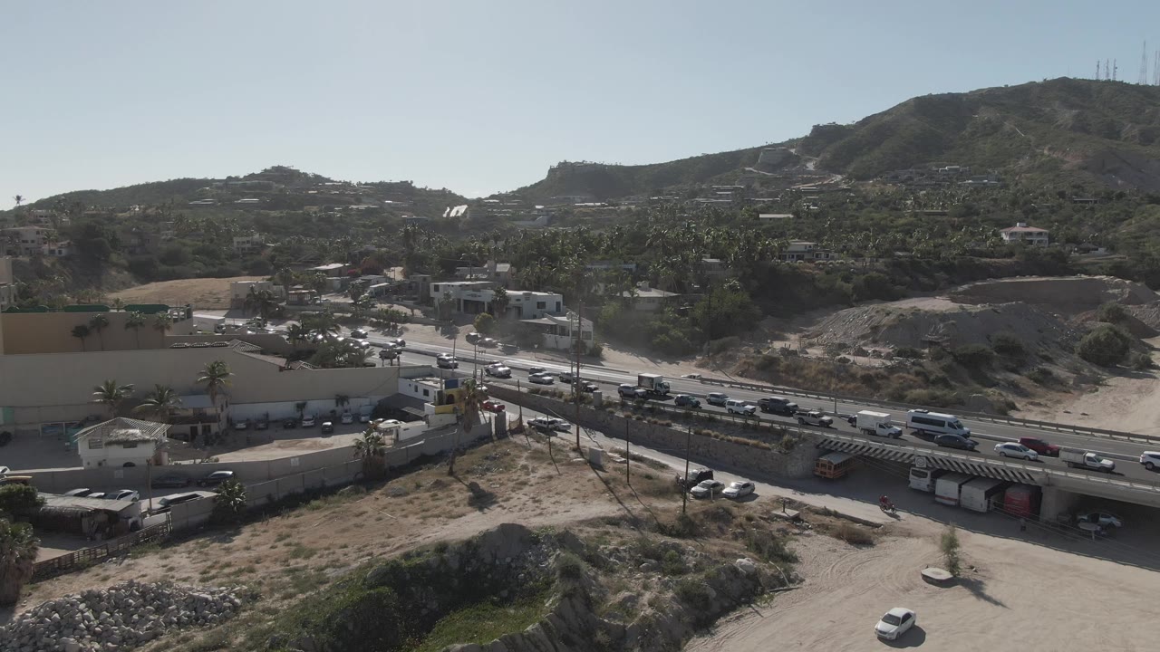 Opening aerial frame of traffic on a multi-lane road in Costa Azul Los Cabos, with a commercial building, parking lot, dry hillside, clear sky, and no visible people.