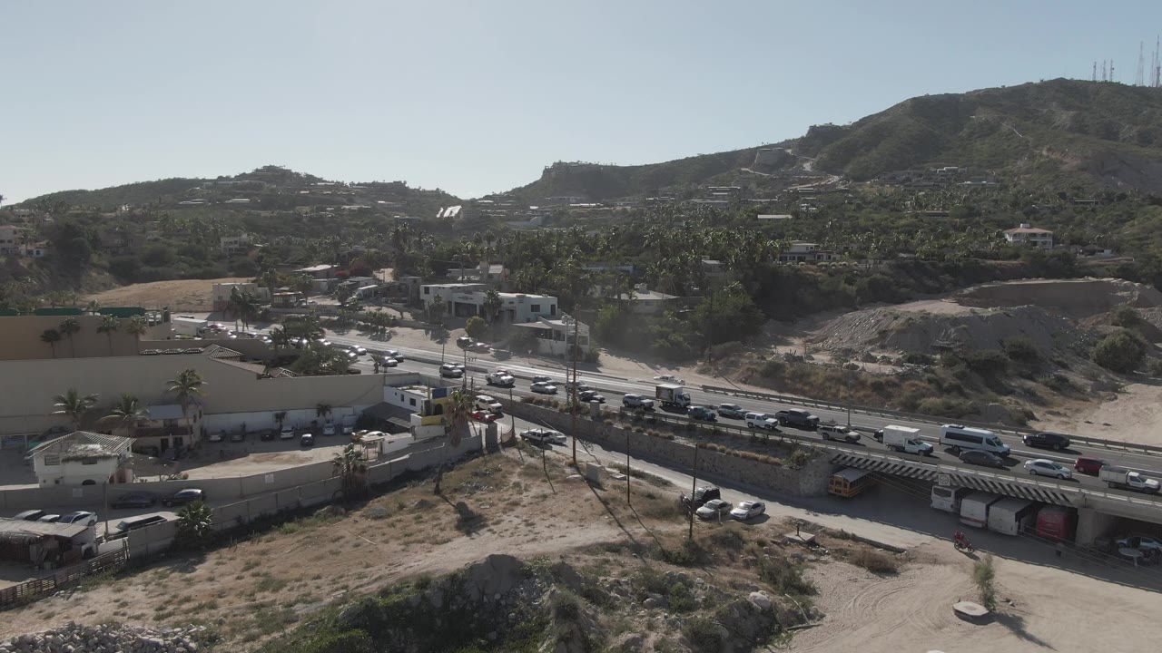 Aerial frame one second in showing vehicles moving along the Costa Azul Los Cabos highway beside a parking area, low buildings, dry terrain, and no visible people.