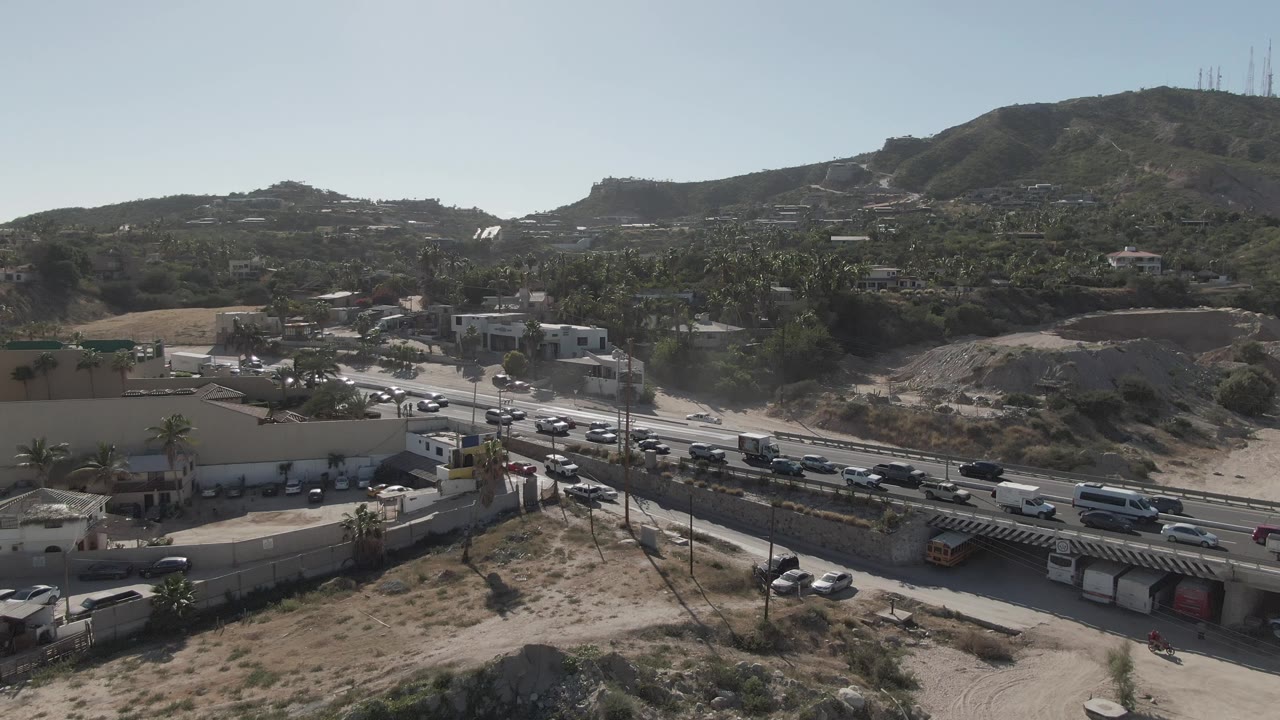 Mid-clip aerial view of Costa Azul Los Cabos traffic, with cars and trucks crossing the frame near a commercial lot, hillside vegetation, distant structures, and no visible people.