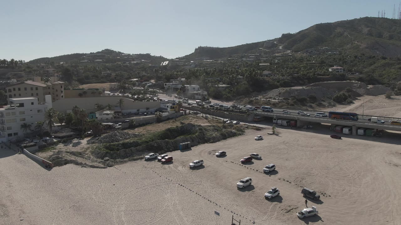 Opening aerial frame over Costa Azul Los Cabos showing a wide dirt parking area, moving road traffic, low buildings, green-brown hills, and no visible people.
