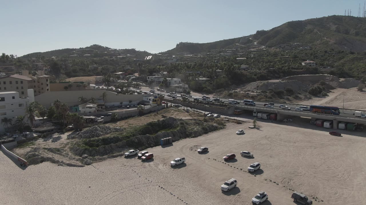 Wide aerial frame with parked cars in a dirt lot and traffic on the road.