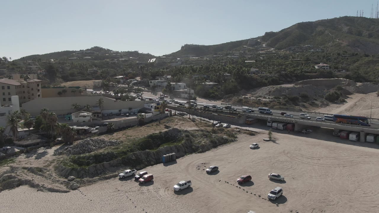 A later aerial frame in Costa Azul Los Cabos shows the dirt lot, clustered parked cars, a truck on the road, hillside homes or buildings, and no visible people.